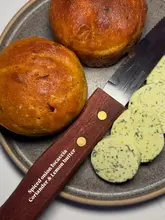 A top-down, close-up shot of two small, golden-brown spiced onion focaccia rolls served alongside slices of coriander and lemon butter on a grey plate. The image features text overlay on the knife handle identifying the dish.