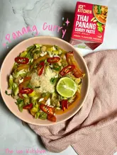 A flat-lay photograph of a homemade Panang curry dish served in a pink bowl, accompanied by the Ice Kitchen curry paste packaging. The image is styled with a pink cloth on a marble surface.