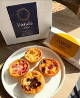 A top-down, natural light photograph featuring four distinct flavours of pastéis de nata arranged on a white plate, flanked by the brand's takeaway packaging.