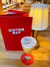 A shot of a cocktail with a foam top, positioned in front of a large, bright red menu and a small table lamp at a bar.