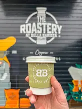 A promotional still image featuring a hand holding a takeaway coffee cup with the 'Bella Barista' logo, positioned in front of the venue's branded roastery wall. The image effectively communicates the brand identity and the coffee-focused nature of the business.