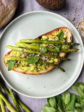 A high-quality, overhead food photography shot featuring a slice of sourdough toast topped with smashed avocado, roasted asparagus, seeds, and fresh mint. The image is bright, well-composed, and clearly highlights the freshness of the ingredients, making it an excellent example of food styling for a recipe or brunch-focused post.