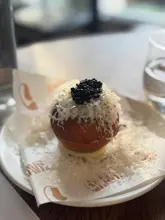 A high-quality, close-up shot of a savory donut topped with caviar and grated cheese, served on branded paper. The image captures the texture and presentation of the dish effectively, using natural light and a shallow depth of field to highlight the food.