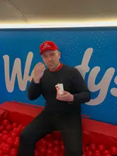 A promotional image featuring a man sitting in a red ball pit at a Wendy's pop-up event, holding a Frosty. The image captures the brand's 'Frosty Funhouse' tour, using the branded backdrop and ball pit to create a playful, casual atmosphere.