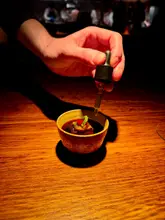 A high-end, atmospheric close-up shot capturing the final touch of a dish being served at a fine dining restaurant. The image features a hand using a dropper to add liquid to a small, elegant bowl, set against a dark, moody background that highlights the craftsmanship of the food.