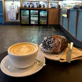 A casual, eye-level shot of a latte with latte art and a chocolate-topped cruffin on a cafe table, with the cafe interior visible in the background.
