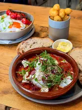 A high-quality, well-lit food photography shot featuring a brunch spread, specifically focusing on a shakshuka dish in the foreground, with a side of hash brown tots and another dish in the background. The composition is clean and inviting, highlighting the textures and freshness of the food.