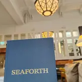 A clean, well-composed photograph of a menu cover for Seaforth restaurant, set against the backdrop of an elegant, well-lit dining room interior. The image effectively uses depth of field to highlight the venue's branding while providing a glimpse of the sophisticated atmosphere.