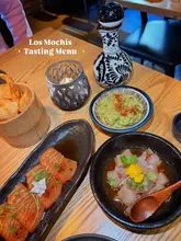 A top-down shot of a tasting menu spread at Los Mochis, featuring salmon tiradito, guacamole with chips, and a ceviche-style dish, illuminated by a candle and a decorative sake bottle.