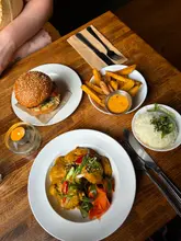A top-down shot of a table spread featuring a gochujang crispy chicken burger, fries with dip, a bowl of rice, and a satay chicken dish, set in a cozy, dimly lit restaurant environment.