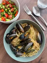 A clean, overhead shot of a bowl of mussels with pasta and a side salad of cucumber and tomato. The lighting is natural and even, and the composition is straightforward and functional.