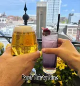 A bright, first-person perspective shot of two people toasting with drinks on a rooftop terrace, with a city skyline visible in the background. The image effectively communicates the venue's atmosphere and setting.