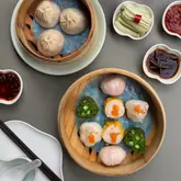 A high-quality, overhead flat-lay photograph of a dim sum spread, featuring bamboo steamers filled with various dumplings, alongside small dishes of dipping sauces and pickled cucumber. The composition is clean and symmetrical, highlighting the vibrant colors and textures of the food against a neutral grey background.