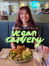 A medium shot of a woman sitting at a restaurant table with a large plate of a vegan Sunday roast in front of her, featuring a prominent green text overlay.