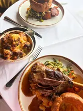 A high-quality, well-lit photograph of a Sunday roast meal, featuring a large portion of braised lamb, a Yorkshire pudding, roasted vegetables, and a side of cauliflower cheese, set on a clean white tablecloth.