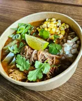 A high-quality, overhead shot of a Birria Ramen bowl featuring slow-braised pulled pork, corn, onions, cilantro, and a lime wedge in a rich, dark broth, presented in a disposable bowl on a wooden surface.
