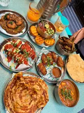 A high-angle, flat-lay shot of a table spread featuring a variety of Indian street food dishes, including skewers, wings, paratha, and curry, accompanied by cocktails.