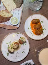 A high-quality, overhead flat-lay photograph of a restaurant table featuring a selection of starters including artisan bread with butter, spiced Red Leicester croquettes, and pulled ham bon bons, alongside cocktails.
