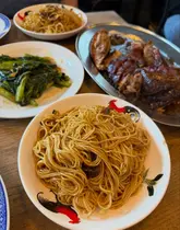 A top-down, overhead shot of a Chinese meal spread on a wooden table, featuring roast duck, wonton noodles, and greens.