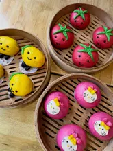 A high-quality, top-down photograph of three bamboo steamers placed on a wooden table, each containing freshly made, character-shaped bao buns designed to look like tomatoes, lemons, and Hello Kitty characters.