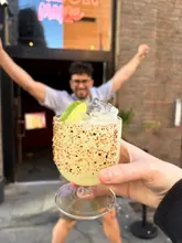 A first-person perspective shot of a cocktail being held up in front of a venue entrance, with a person celebrating in the background.