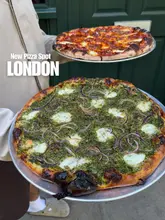 A casual, eye-level shot of two pizzas being held in front of a green door, likely outside a restaurant. The pizzas appear to be a pepperoni option and a green-topped vegetable option.