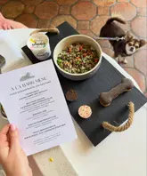 A high-quality, well-composed photograph showcasing a 'doggy afternoon tea' experience at a hospitality venue. The image features a menu titled 'A La Bark,' a bowl of fresh food, and dog-friendly treats arranged on a slate board, with a dog visible in the background. The lighting is bright and natural, and the presentation is charming and on-brand for a pet-friendly establishment.