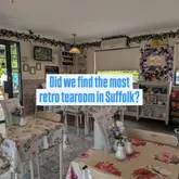 A wide-angle shot of a retro-themed tea room in Suffolk, showcasing the venue's interior with floral tablecloths, artificial flower arrangements, and vintage-style crockery. The image serves as a venue tour to highlight the nostalgic atmosphere.