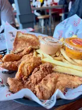 A close-up, eye-level shot of a fast-food meal consisting of breaded chicken tenders, fries, a slice of toast, and dipping sauces in a plastic basket, typical of a casual dining experience.
