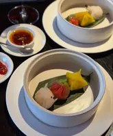 A high-quality, top-down shot of a dim sum course served in a bamboo steamer, featuring colorful dumplings garnished with caviar. The setting is elegant, with small dipping sauce bowls and a wine glass visible, suggesting a sophisticated dining experience at a hotel restaurant.