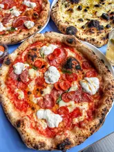 A high-angle, close-up shot of Neapolitan-style pizzas on a blue table. The focus is on a pepperoni pizza topped with dollops of stracciatella, with two other pizzas partially visible in the background and a cocktail glass on the side.