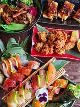 A high-angle flat lay photograph of a Japanese dining spread featuring a sushi platter, roast duck mazeudon, chicken karaage, and truffle teriyaki tofu arranged on a wooden table.