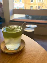 A high-quality, well-lit photograph of a layered coconut matcha drink served in a glass on a wooden coaster, set in a bright, modern cafe environment with a blurred window view in the background.