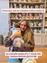 A lifestyle photograph featuring a mother holding her newborn baby in a pottery cafe, capturing a 'first day out' moment. The setting is filled with shelves of ceramics, creating a cozy, domestic atmosphere.