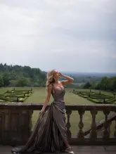 A high-quality, atmospheric shot of a woman in an elegant evening gown standing on the terrace of a historic estate, likely Cliveden House. The image captures a sense of luxury and occasion, focusing on the grandeur of the setting and the fashion.