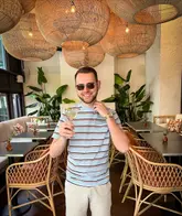 A man posing with a cocktail in a stylish, well-lit bar setting with distinctive wicker lighting, promoting a new cocktail menu at a hotel bar.