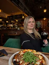 A lifestyle-style shot featuring a person holding a glass of red wine in a restaurant setting, with a pizza in the foreground. The image aims to capture the atmosphere of the venue, showing the dining area and the food.