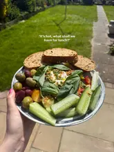 A top-down, hand-held shot of a homemade burrata and salad plate featuring olives, cucumber, and bread, taken outdoors in a garden setting.