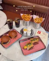 A bright, overhead flat-lay shot of a meal at Zula Burger, featuring two burgers, a hot dog, two portions of fries, and drinks served on branded trays on a marble table.