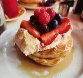 A close-up, angled shot of a stack of pancakes topped with vanilla cream, fresh strawberries, raspberries, and blueberries, drizzled with maple syrup at The Breakfast Club.