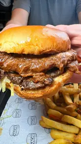 A creator reviews a burger restaurant in Manchester, showcasing several menu items including smash burgers with brisket, chicken tenders, and loaded fries. The video also highlights the venue's 'bunker' games room and film club, positioning it as a great spot for parties and social gatherings.