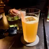 A photo of a cocktail in a tall glass with a large ice cube, taken in a dimly lit bar setting.