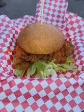 A close-up, eye-level shot of a fried chicken burger served in a red and white checkered basket, typical of a street food setting.