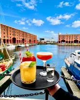 A POV shot of a tray of cocktails held up against the backdrop of the Albert Dock in Liverpool. The image is heavily saturated and sharpened, creating a vibrant but somewhat artificial aesthetic.