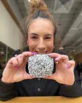 A close-up portrait of a creator holding a gluten-free and vegan lamington-style doughnut in front of their face. The image is a direct product showcase, likely taken in a retail environment.