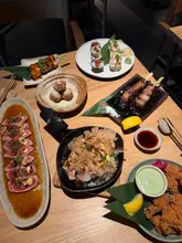 A high-quality, overhead flat-lay photograph of a Japanese feast at Sticks'n'Sushi. The table is filled with a variety of dishes including sushi, skewers, and small plates, all arranged in an aesthetically pleasing manner that highlights the restaurant's presentation style.