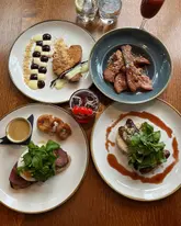 A high-quality, overhead flat-lay photograph of a brunch spread featuring four distinct plates and a cocktail, set on a wooden table. The dishes include steak and eggs, a loaded toast dish, sausage and crumpets, and pancakes with bacon, all presented with care and good lighting.