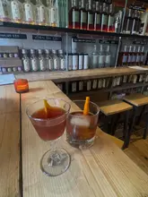 A high-quality, eye-level shot of two cocktails served on a wooden bar counter. The background features a well-stocked bar shelf with various spirit bottles and handwritten price labels, creating an authentic and atmospheric setting for a cocktail bar.