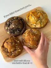 A high-quality, well-lit shot of four thick, gourmet-style cookies arranged on a wooden board, with a hand reaching in to grab one, effectively showcasing the texture and variety of the product.