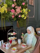 A promotional image featuring a woman enjoying a themed afternoon tea at The Clermont, London. The setting is elegant with a large floral display, showcasing a traditional three-tier stand with sandwiches, scones, and pastries.